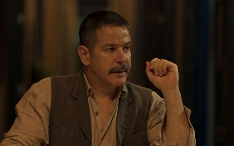 Tenório (Murilo Benício) está sentado em mesa e tem expressão de descontentamento em cena de Pantanal, novela das nove da Globo