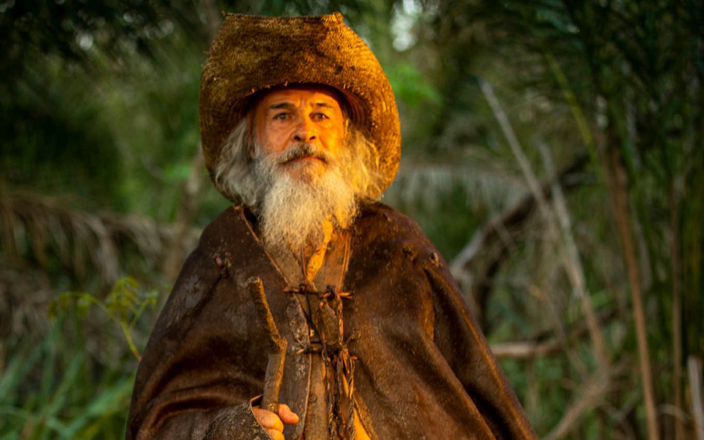 O ator Osmar Prado com longas barbas e cabelos brancos com uma capa preta envelhecida como o Velho do Rio em cena de Pantanal