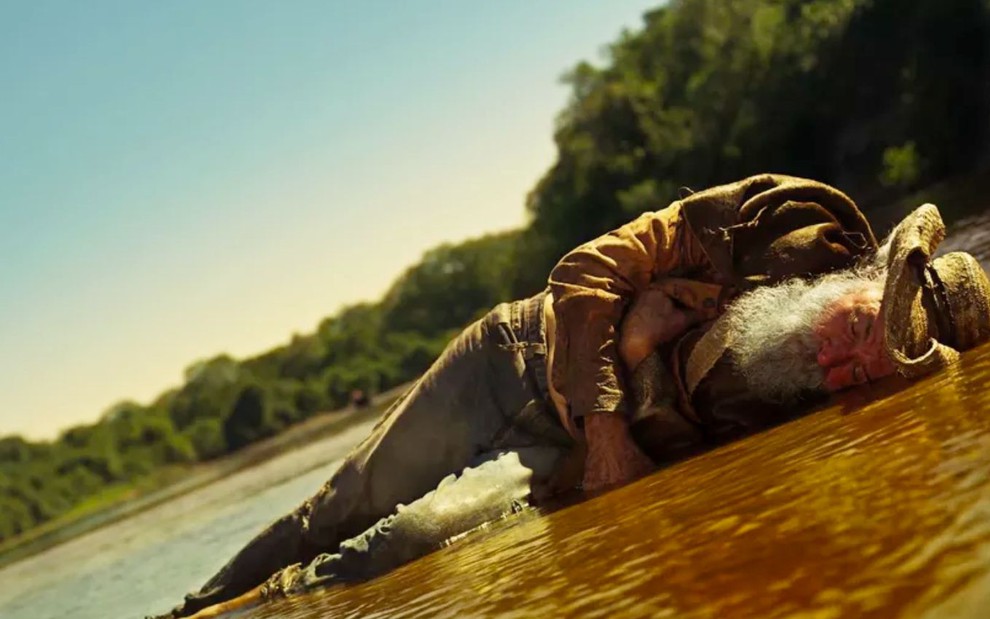 O ator Osmar Prado como Velho do Rio em Pantanal; ele está caído à beira do rio após levar um tiro