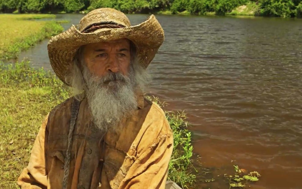 O ator Osmar Prado sentado em uma canoa como o Velho do Rio em cena de Pantanal
