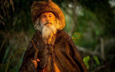 Osmar Prado caracterizado como Velho do Rio. Ele usa o cabelo e a barba longas, e veste chapéu de palha e casaco de couro