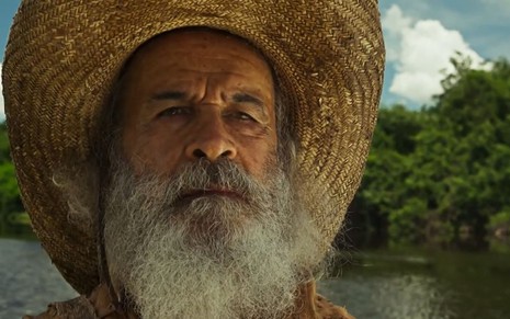 Velho do Rio (Osmar Prado) está sentado em barco em cena de Pantanal; ele usa chapéu de palha
