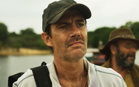Rafael Sieg tem a barba malfeita e usa um boné verde; ele dá um sorriso maligno em cena de Pantanal