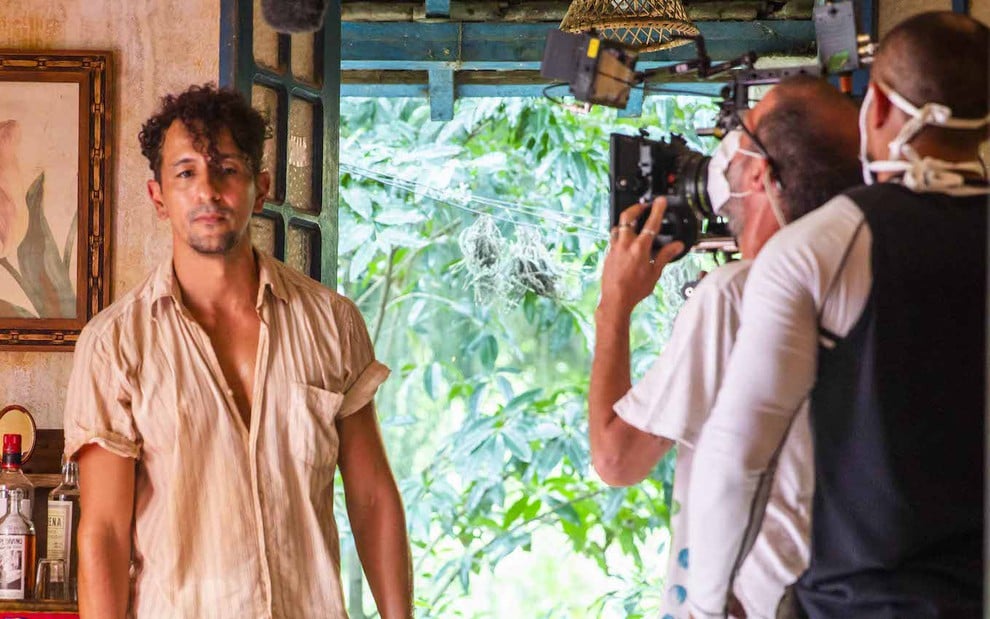 O ator Irandhir Santos exibe cabelo cacheado e camisa semiaberta ao ser acompanhado por técnicos em gravação de cena de Pantanal