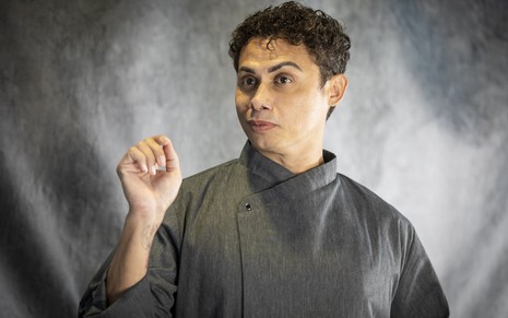O ator Silvero Pereira está com um uniforme cinza de mordomo, com um dos braços levantados, caracterizado como Zaquieu da novela Pantanal