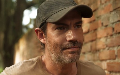 Rafael Sieg tem a barba malfeita e usa um boné verde; ele dá um sorriso sinistro em cena de Pantanal
