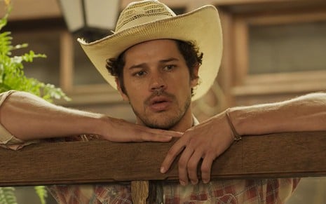 José Loreto caracterizado como Tadeu em Pantanal; ator está escorado em um painel de madeira, com a expressão séria