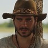 Gabriel Sater caracterizado como Trindade de Pantanal: ele veste chapéu de palha e camisa surrada, e exibe uma expressão séria em cena de Pantanal