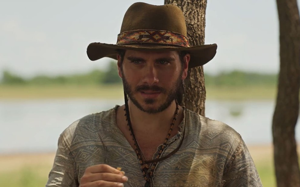 Gabriel Sater caracterizado como Trindade de Pantanal: ele veste chapéu de palha e camisa surrada, e exibe uma expressão séria em cena de Pantanal