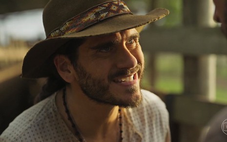 O ator e cantor Gabriel Sater sorri de maneira sinistra em cena de Pantanal na qual usa um chapéu e está caracterizado como Trindade