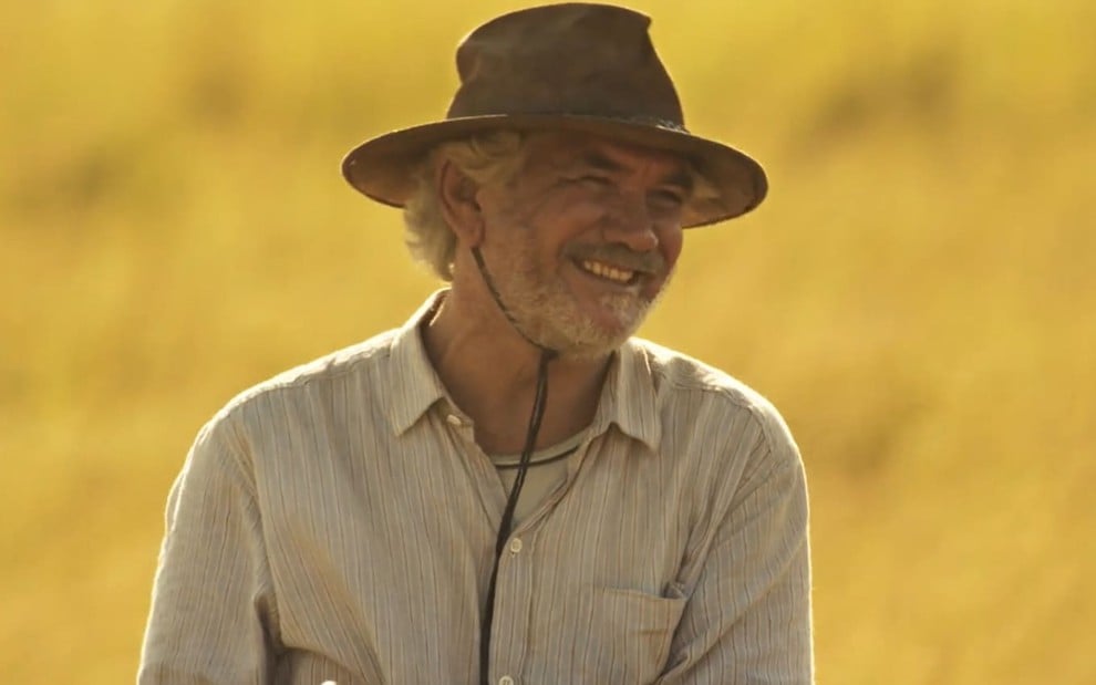 Jackson Antunes caracterizado como Túlio: ator veste uma camiseta surrada e um chapéu. Ele dá um leve sorriso em cena de Pantanal.