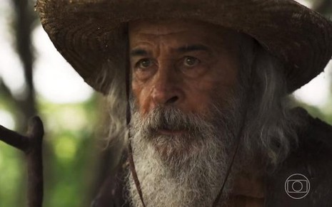 O ator Osmar Prado segura cajado e usa um chapéu em cena de Pantanal na qual está caracterizado como o Velho do Rio