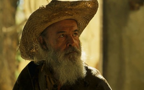 Osmar Prado caracterizado como Velho do Rio. Ele usa o cabelo e a barba longas e veste chapéu de palha e capa de couro. A expressão está séria em cena de Pantanal