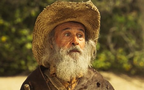 Osmar Prado caracterizado como Velho do Rio. Ele usa o cabelo e a barba longas, e veste chapéu de palha e casaco de couro e tem a expressão debochada em cena de Pantanal