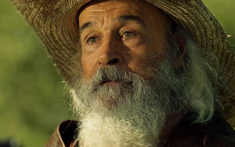 Osmar Prado caracterizado como Velho do Rio. Ele tem a barba longa e veste chapéu de palha e casaco de pele. A expressão do ator está emocionada