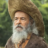 O ator Osmar Prado segura cajado, usa capa surrada e um chapéu em cena de Pantanal na qual está caracterizado como o Velho do Rio