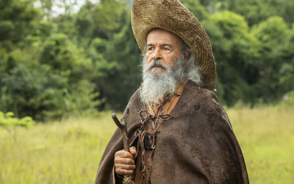 O ator Osmar Prado segura cajado, usa capa surrada e um chapéu em cena de Pantanal na qual está caracterizado como o Velho do Rio