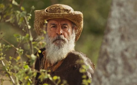 Osmar Prado estpa com chapéu atrás de um arbusto em cena de Pantanal como o Velho do Rio