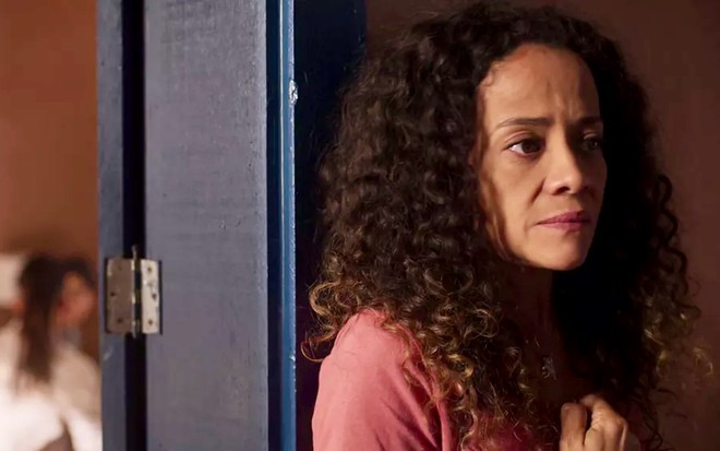 A atriz Aline Borges está encostada no batende de uma porta com uma expressão desagradável em cena de Pantanal