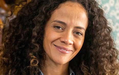 Aline Borges caracterizada como Zuleica: atriz usa um vestido jeans e dá um leve sorriso; ela tem os cabelos cacheados e cheios em ensaio de divulgação de Pantanal