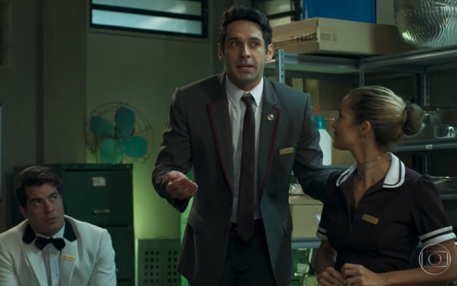 João Baldasserini grava de uniforme de recepcionista ao lado de Nanda Costa, de costas na foto, como Agnaldo e Sandra Helena