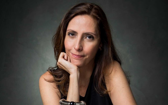 A autora Claudia Solto leva a mão direito ao queixo em que apoia a cabeça enquanto olha diretamente para a câmera