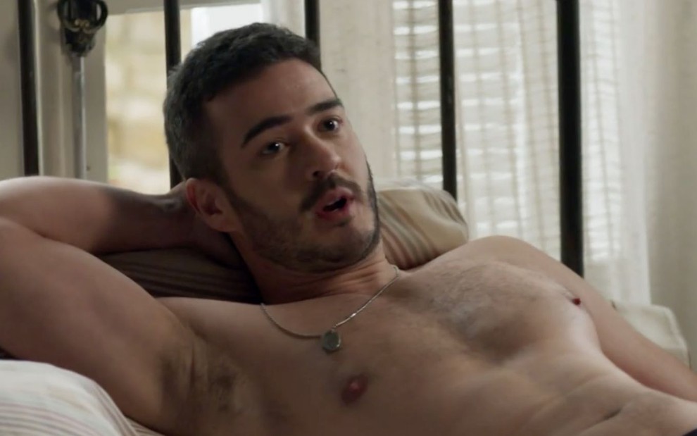 Marcos Veras em cena de Pega Pega: ator está deitado na cama, sem camisa e olha para alguém fora do quadro