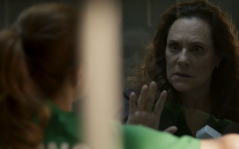 Arlete (Elizabeth Savala) está sentada em sala de visita em cadeia, ela está com a mão no vidro fazendo um sinal para Mônica (Julia Lund), que está de costas na foto, esperar em cena de Pega Pega