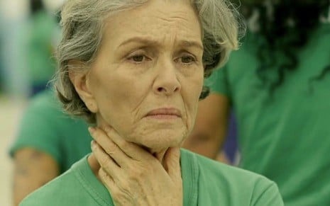 A atriz Irene Ravache como Sabine de cara limpa, com a mão direita no pescoço, enquanto usa uma camisa verde do sistema prisional em cena de Pega Pega