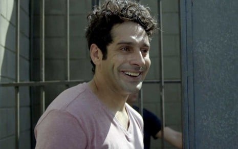 O ator João Baldasserini caracterizado como Agnaldo está dentro de uma cadeia, com uma cela às suas costas em cena de Pega Pega