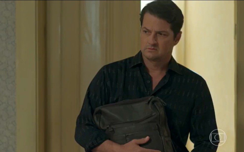 Malagueta (Marcelo Serrado) com uma bolsa de couro na mão; ele veste camisa social preta