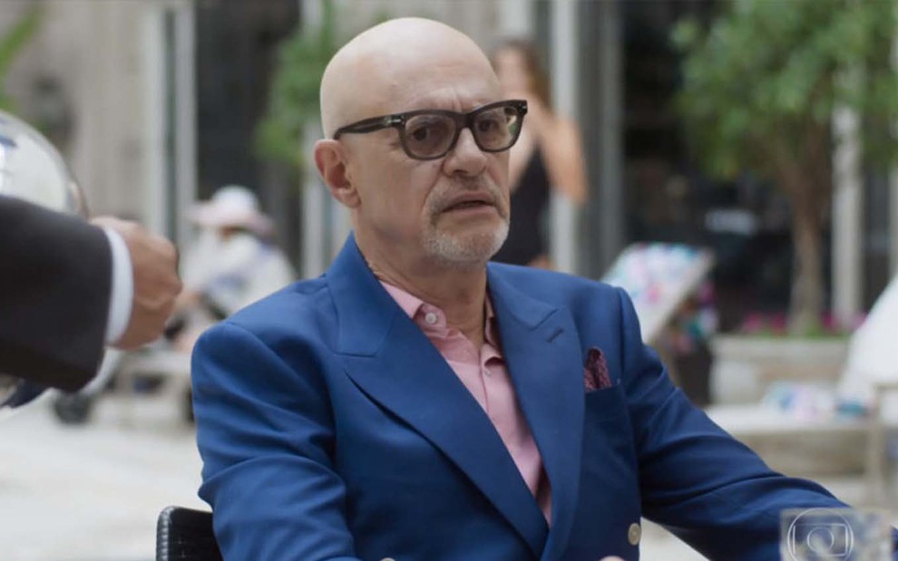 O ator Marcos Caruso está sentado à mesa na área externa de um hotel com um blazer azul e uma camisa rosa como Pedrinho Guimarães em Pega Pega