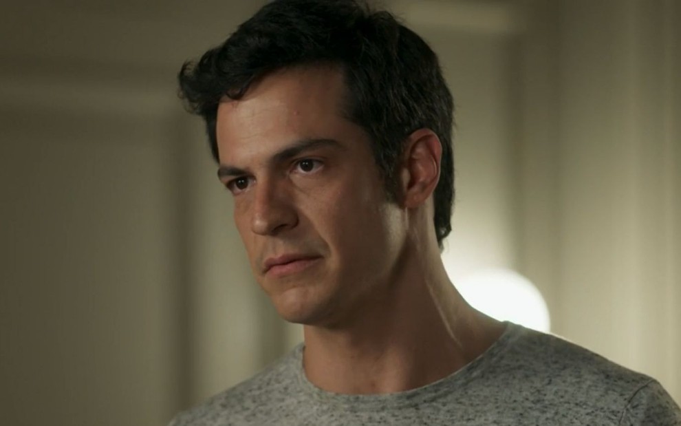 Eric (Mateus Solano) está em pé na sala de Lourenço (Romulo Arantes Neto); ele usa uma camiseta cinza em cena de Pega Pega