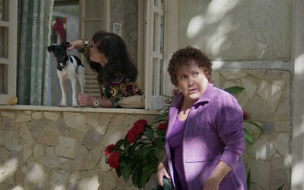 A atriz Cristina Pereira como Prazeres está na janela e faz carinho em um cachorro, enquanto Nicette Bruno está na frente da casa como Elza em cena de Pega Pega