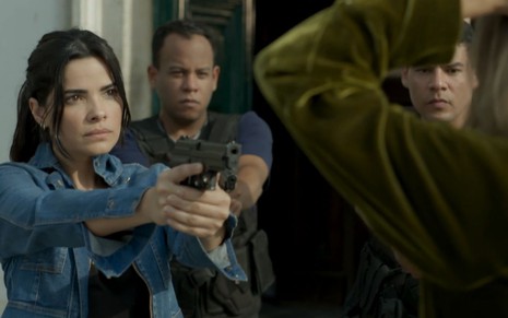 Vanessa Giacomo grava apontando arma em direção à Nanda Costa, que aparece de costas e com os braços na cabeça na imagem, como Antônia e Sandra Helena em Pega Pega