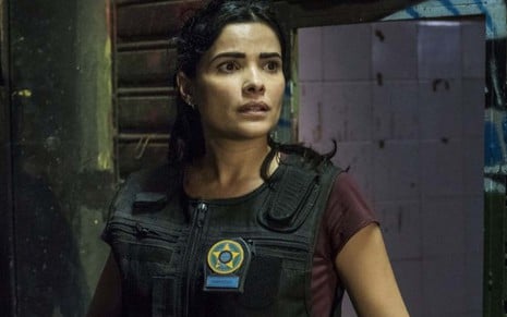 A atriz Vanessa Giácomo está em um beco escuro como Antônia com um colete à prova de balas e um distintivo da Polícia Civil do lado direito do peito em cena de Pega Pega