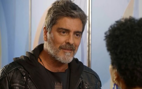 Junno Andrade, caracterizado como Renato, em cena de Poliana Moça; ele dá um leve sorriso enquanto encara Elina de Souza, a Helô