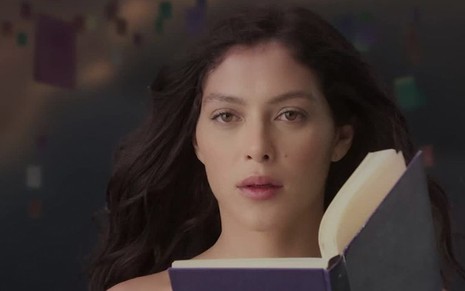 A atriz A Maia, em close-up, como a Morte segurando um livro em cena de Quanto Mais Vida, Melhor