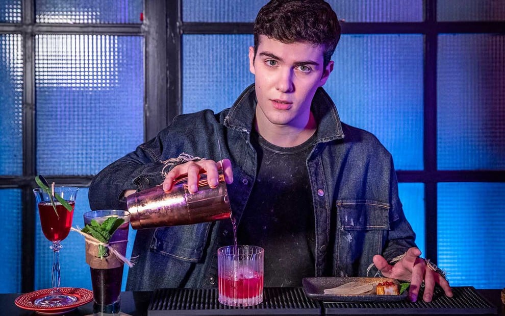 Em um bar, o ator Caio Manhente como Gabriel vira um drink rosa em um copo com gelo em cena de Quanto Mais Vida, Melhor!