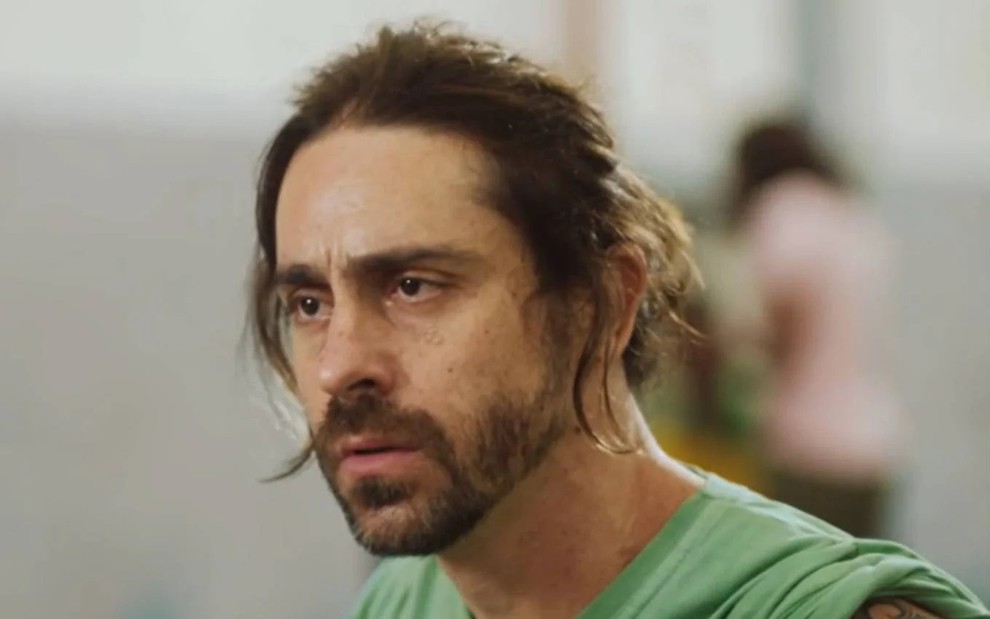 Felipe Abib, caracterizado como Roni, tem a expressão perturbada em Quanto Mais Vida, Melhor!; ator tem os cabelos presos e usa uma blusa verde