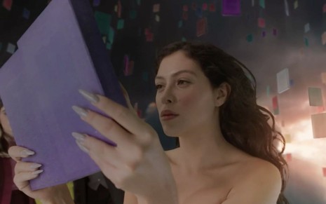 A Maia em cena de Quanto Mais Vida, Melhor!: desnuda, atriz segura livro de capa lilás e está com os cabelos esvoaçantes