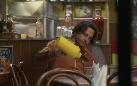 O ator Vladimir Brichta como Neném sentado a mesa em um bar virando uma garrafa de cerveja em um copo em cena de Quanto Mais Vida, Melhor