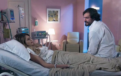 A atriz Bruna Gerin, deitada em uma cama de hospital, sendo observada pelo ator Thiago Lacerda, em cena de Salve-se Quem Puder