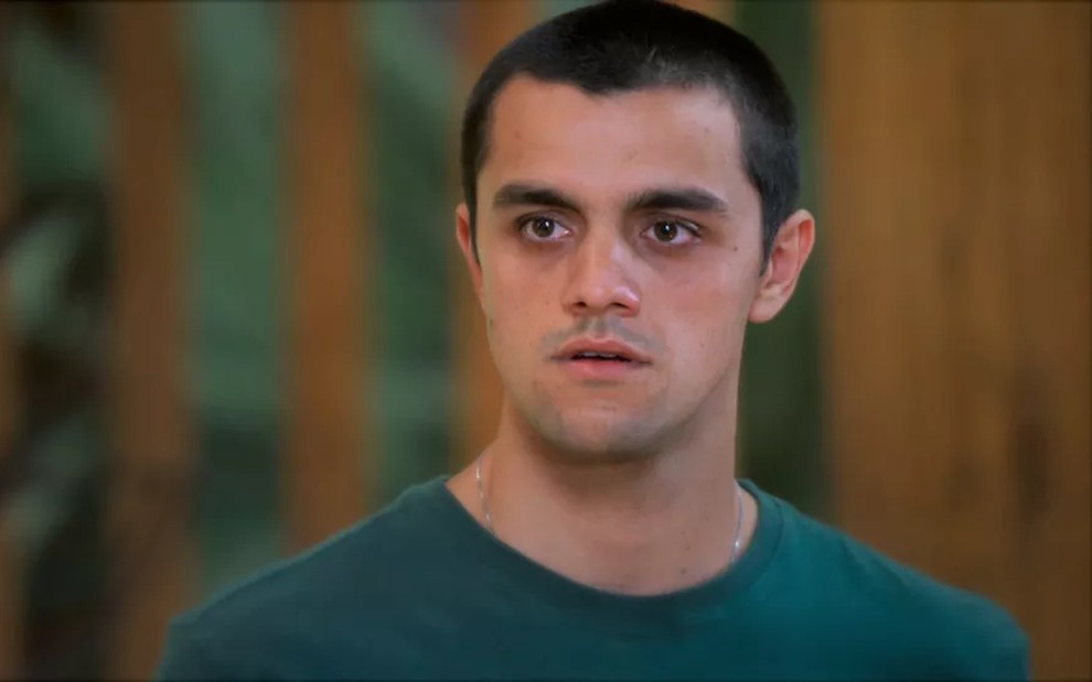 O ator Felipe Simas, com uma camiseta verde e expressão de espanto, em cena como Téo em Salve-se Quem Puder