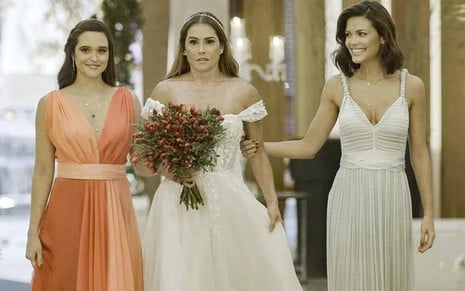 As atrizes Juliana Paiva e Vitória Strada usam vestidos de festas; Deborah Secco, no meio, está vestida de noiva em cena de casamento em Salve-se Quem Puder