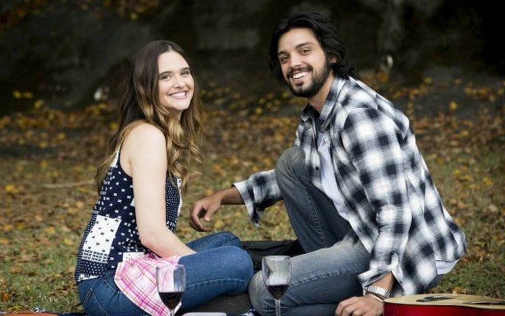 Os atores Juliana Paiva e Rodrigo Simas, sentados em um gramado, em cena como Luna e Alejandro em Salve-se Quem Puder