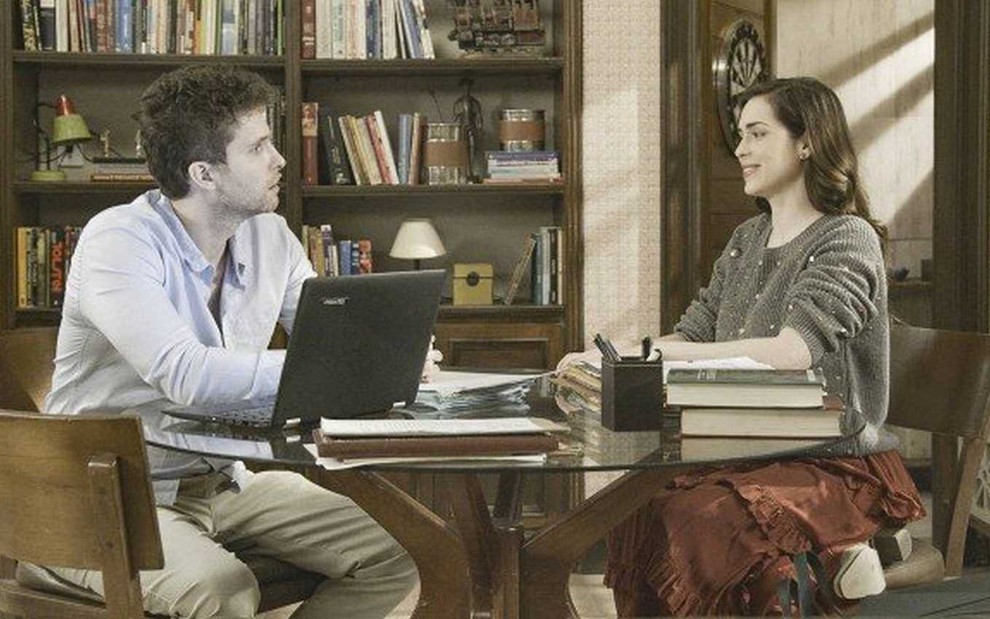 Os atores Thiago Fragoso e Sophia Abrahão, sentados em uma mesa de trabalho, em cena como Alan e Júlia em Salve-se Quem Puder