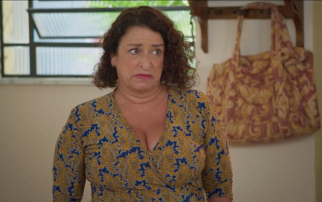 A atriz Grace Gianoukas com expressão de tristeza como Ermelinda, ela está em frente a uma parede que tem uma báscula entreaberta e um cabideiro com uma bolsa amarela com flores rosas pendurada em cena de Salve-se Quem Puder