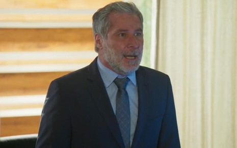 O ator Leopoldo Pacheco está em uma sala de reunião, de terno azul marinho, blusa azul e gravata preta, como Hugo em cena de Salve-se Quem Puder