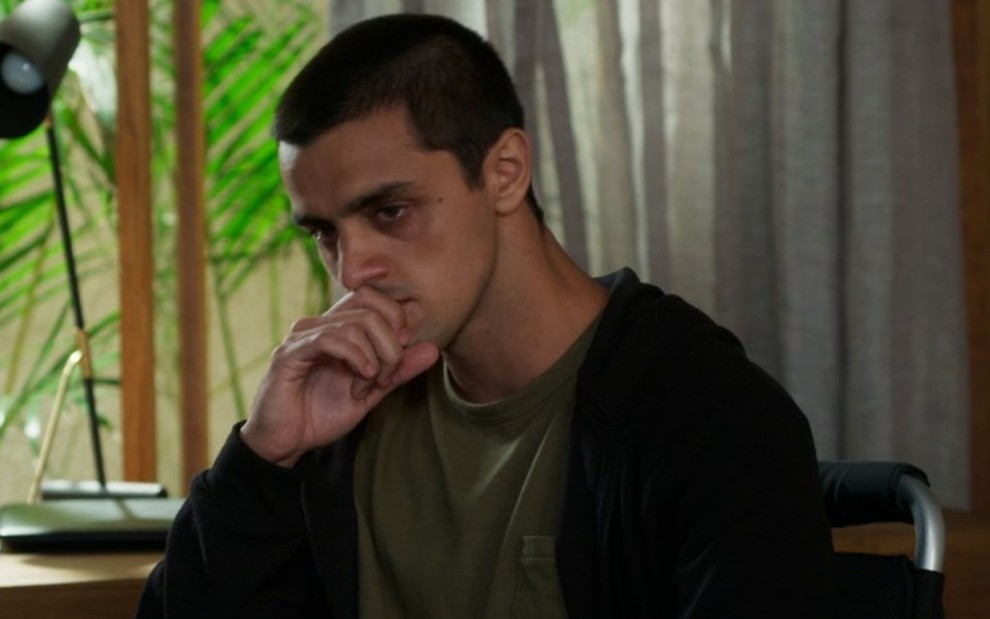 Felipe Simas caracterizado como Téo em cena de Salve-se Quem Puder: ator está sentado e com a mão apoiada no rosto em sinal de tristeza